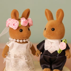 May include: Two brown rabbit figurines dressed for a wedding. One rabbit wears a white dress, veil, and pearl necklace, and a pink rose flower crown. The other rabbit wears a black tuxedo with a white bow tie and a pink rose boutonniere.