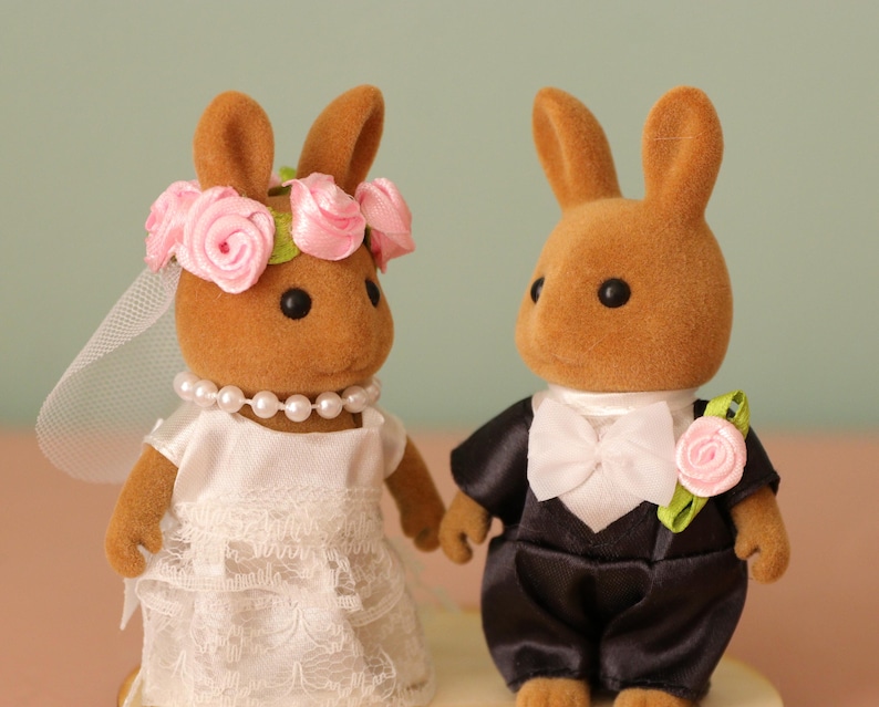 May include: Two brown rabbit figurines dressed for a wedding. One rabbit wears a white dress, veil, and pearl necklace, and a pink rose flower crown. The other rabbit wears a black tuxedo with a white bow tie and a pink rose boutonniere.