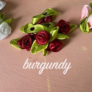 May include: Three clusters of small ribbon roses with green leaves, arranged in groups labeled white, burgundy, and pink. Each cluster consists of multiple fabric flowers crafted from satin ribbon, featuring distinct color variations for each group.