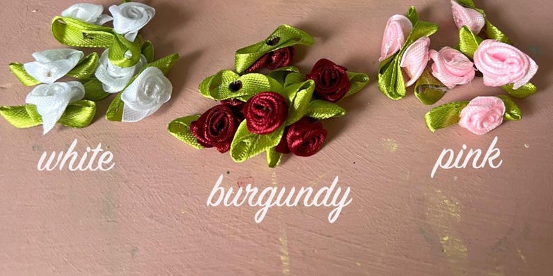 May include: Three clusters of small ribbon roses with green leaves, arranged in groups labeled white, burgundy, and pink. Each cluster consists of multiple fabric flowers crafted from satin ribbon, featuring distinct color variations for each group.