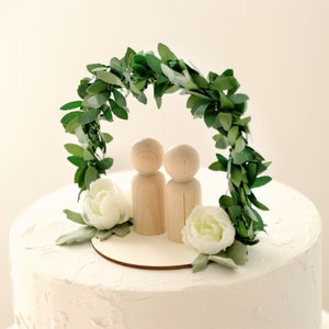 Puede incluir: Adorno para tarta de boda con dos mu&ntilde;ecos de madera bajo un arco de hojas verdes. Flores blancas y una base de madera en la base del arco. La tarta tiene glaseado blanco.