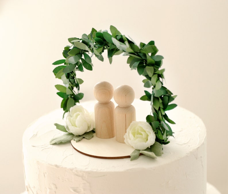 Puede incluir: Adorno para tarta de boda con dos mu&ntilde;ecos de madera bajo un arco de hojas verdes. Flores blancas y una base de madera en la base del arco. La tarta tiene glaseado blanco.