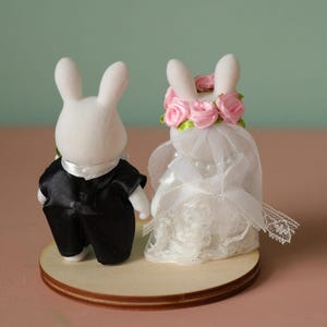 May include: Two white flocked rabbit figurines standing on a circular wooden base. The left rabbit wears a black satin tuxedo jacket with a white collar. The right rabbit wears a white lace dress, a pearl necklace, and a pink rose floral crown.