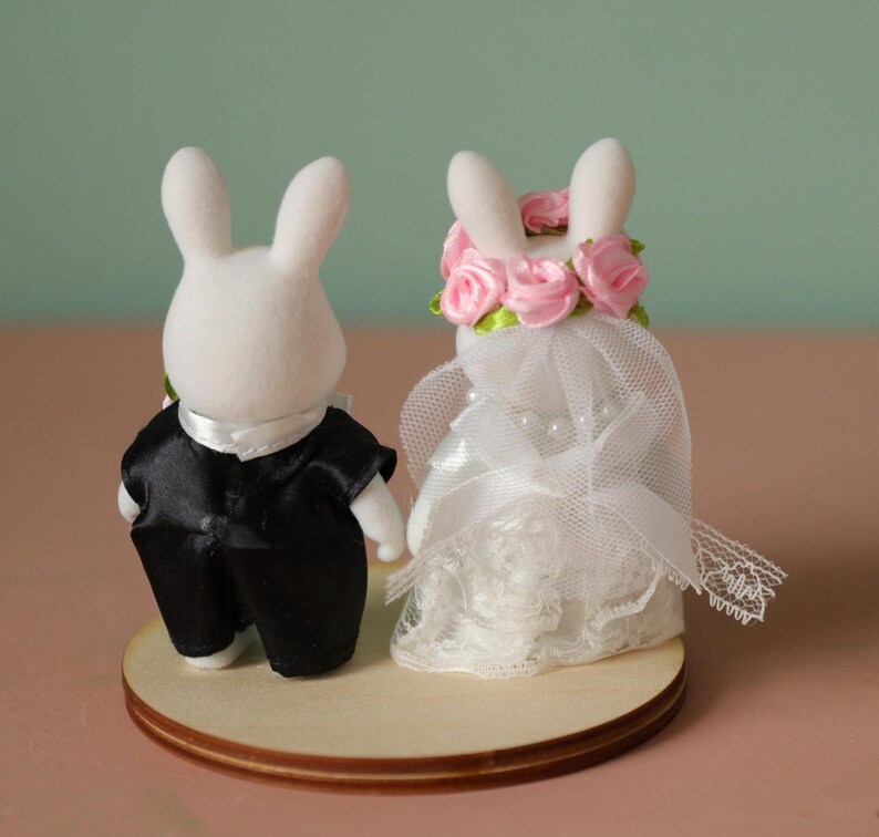 May include: Two white flocked rabbit figurines standing on a circular wooden base. The left rabbit wears a black satin tuxedo jacket with a white collar. The right rabbit wears a white lace dress, a pearl necklace, and a pink rose floral crown.