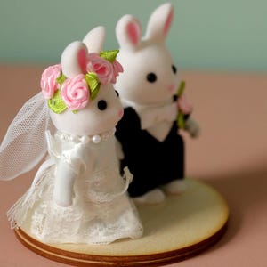 May include: Two white flocked rabbit figurines standing on a circular wooden base. The left rabbit wears a white lace dress, a pearl necklace, and a floral crown with pink roses and green leaves. The right rabbit wears a black tuxedo with a white bow tie.