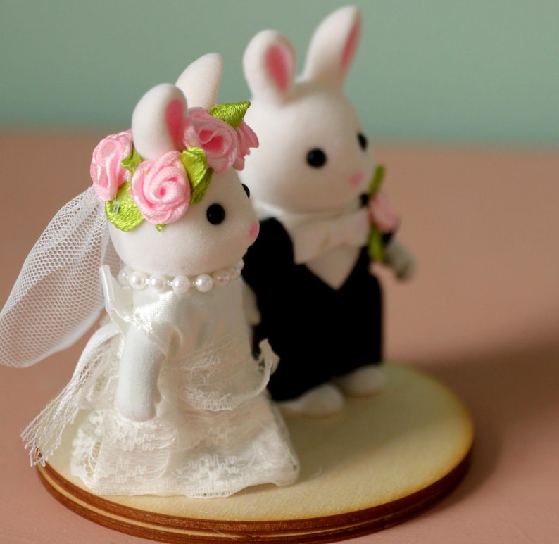 May include: Two white flocked rabbit figurines standing on a circular wooden base. The left rabbit wears a white lace dress, a pearl necklace, and a floral crown with pink roses and green leaves. The right rabbit wears a black tuxedo with a white bow tie.