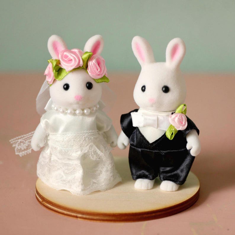May include: Two white flocked rabbit figurines dressed for a wedding. The left rabbit wears a white satin dress with lace trim, a pearl necklace, and a floral crown with pink roses and green leaves. The right rabbit wears a black tuxedo with a white bow tie and a pink rose boutonniere.