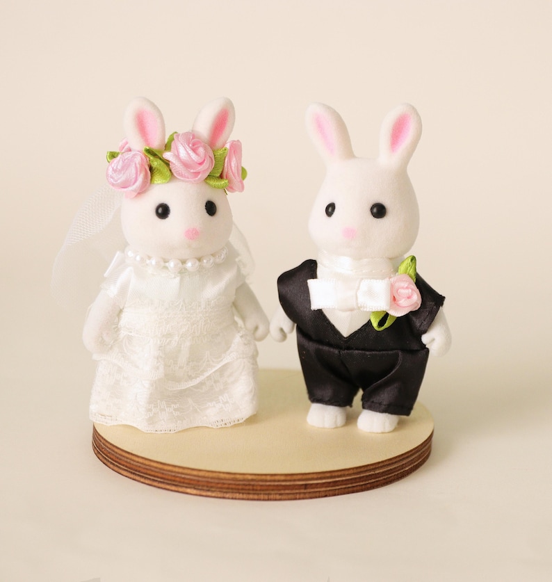 May include: Two white rabbit figurines standing on a circular wooden base. The left rabbit wears a white lace dress, a pearl necklace, and a pink rose floral crown with a veil. The right rabbit wears a black tuxedo jacket with a white bow tie.