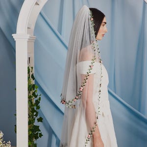 Peut inclure: Un voile de mariage blanc avec une délicate bordure florale de roses roses et de feuilles vertes. Le voile transparent est porté avec une robe de mariée blanche à épaules dénudées. L'arrière-plan présente une arche blanche et du tissu bleu.