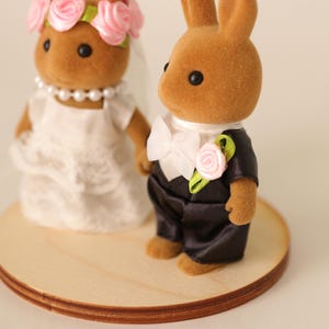 May include: A wedding cake topper featuring two brown rabbit figurines. The bride rabbit wears a white dress, veil, and a pink rose crown. The groom rabbit wears a black tuxedo with a white bow tie and a pink rose boutonniere. The figurines stand on a round wooden base.