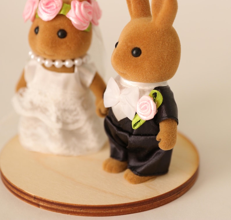 May include: A wedding cake topper featuring two brown rabbit figurines. The bride rabbit wears a white dress, veil, and a pink rose crown. The groom rabbit wears a black tuxedo with a white bow tie and a pink rose boutonniere. The figurines stand on a round wooden base.