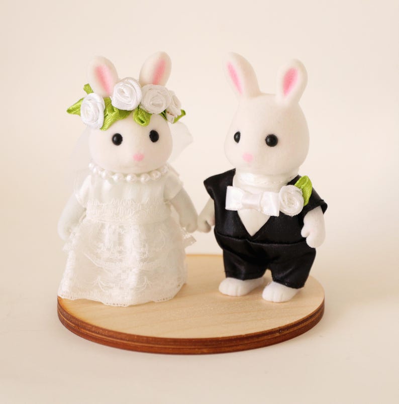 May include: Two white flocked rabbit figurines standing on a circular wooden base. The left rabbit wears a white lace dress, a pearl necklace, and a floral crown. The right rabbit wears a black tuxedo jacket with a white bow tie and a flower.