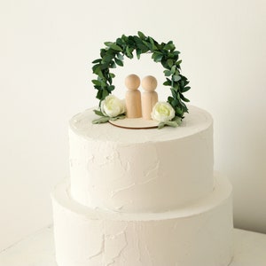 Puede incluir: Una tarta de boda blanca de dos pisos, coronada con un adorno de tarta de madera. El adorno presenta dos figuras de madera bajo un arco de hojas verdes, con flores blancas y vegetaci&oacute;n. La tarta tiene una cobertura de crema de mantequilla texturizada.