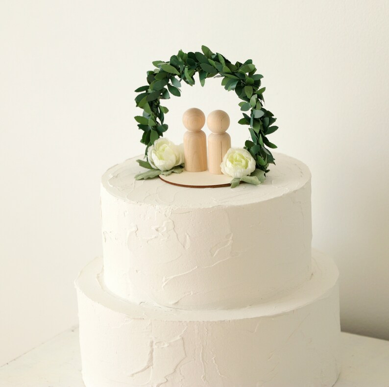 Puede incluir: Una tarta de boda blanca de dos pisos, coronada con un adorno de tarta de madera. El adorno presenta dos figuras de madera bajo un arco de hojas verdes, con flores blancas y vegetaci&oacute;n. La tarta tiene una cobertura de crema de mantequilla texturizada.