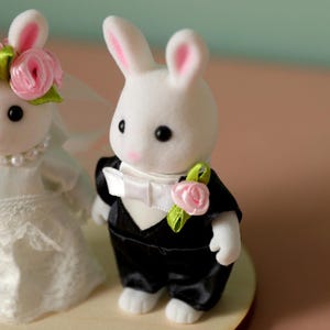 May include: Two white flocked rabbit figurines dressed for a wedding. One rabbit wears a white lace dress with a pearl necklace and pink rose headpiece. The other rabbit wears a black tuxedo jacket with a white bow tie and a pink rose boutonniere.