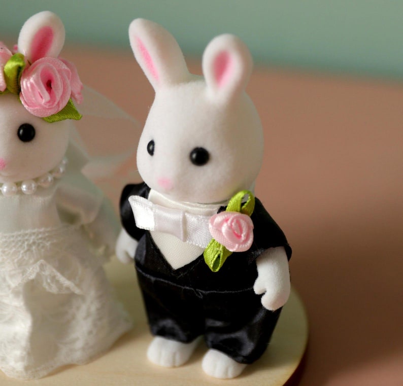 May include: Two white flocked rabbit figurines dressed for a wedding. One rabbit wears a white lace dress with a pearl necklace and pink rose headpiece. The other rabbit wears a black tuxedo jacket with a white bow tie and a pink rose boutonniere.