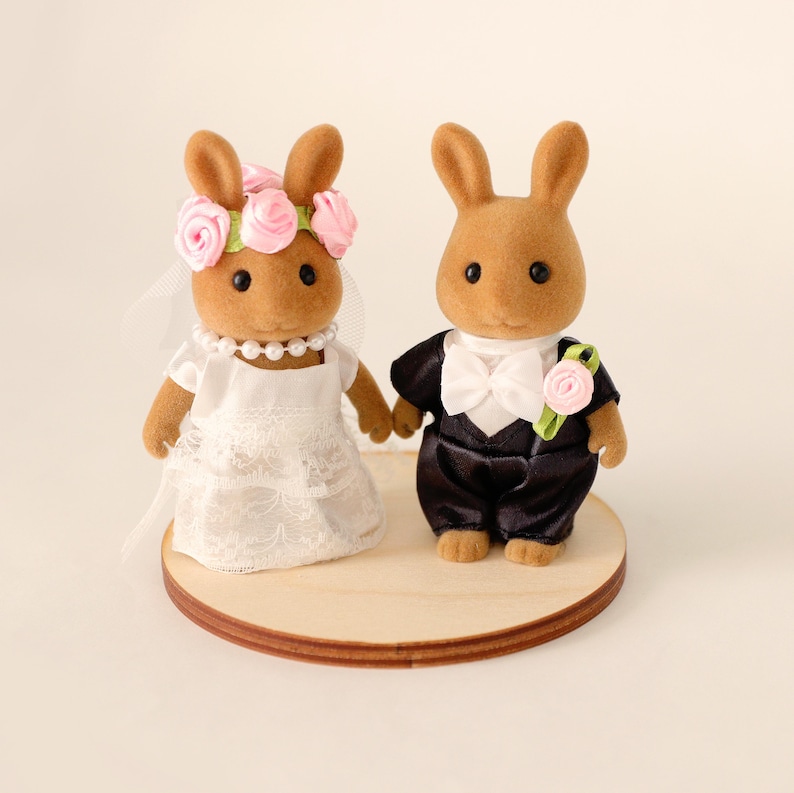 May include: Two brown rabbit figurines dressed for a wedding. The bride rabbit wears a white lace dress, pearl necklace, and a pink rose flower crown. The groom rabbit wears a black tuxedo with a white bow tie and a pink rose boutonniere. They stand on a wooden base.
