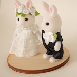May include: Two white flocked rabbit figurines dressed for a wedding. The left rabbit wears a white lace dress, a pearl necklace, and a floral headpiece. The right rabbit wears a black tuxedo with a white bow tie and a small green leaf lapel pin.