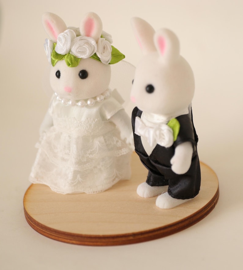 May include: Two white flocked rabbit figurines dressed for a wedding. The left rabbit wears a white lace dress, a pearl necklace, and a floral headpiece. The right rabbit wears a black tuxedo with a white bow tie and a small green leaf lapel pin.