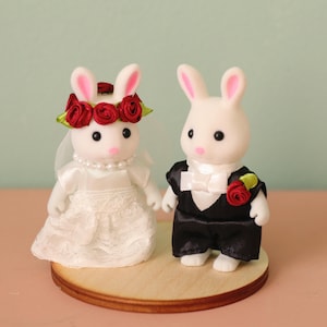 May include: Two white flocked rabbit figurines dressed for a wedding. The left rabbit wears a white lace dress, a pearl necklace, and a red rose crown with a veil. The right rabbit wears a black tuxedo jacket, white shirt, bow tie, and a red rose boutonniere.