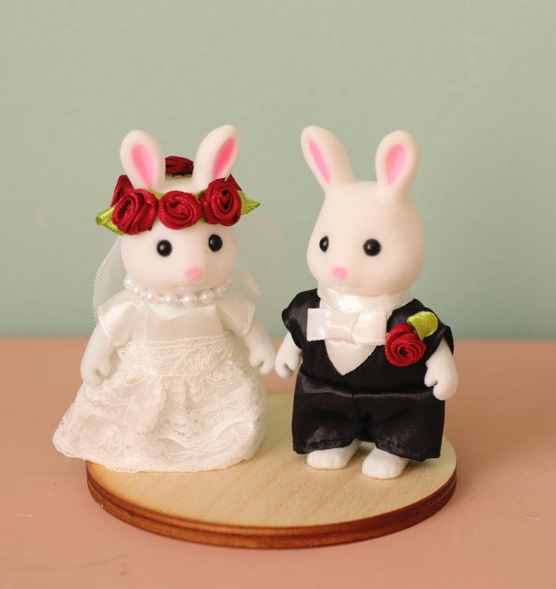 May include: Two white flocked rabbit figurines dressed for a wedding. The left rabbit wears a white lace dress, a pearl necklace, and a red rose crown with a veil. The right rabbit wears a black tuxedo jacket, white shirt, bow tie, and a red rose boutonniere.