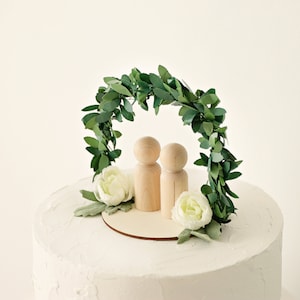 Puede incluir: Un adorno para tarta de boda con dos mu&ntilde;ecos de madera sobre una base de madera, bajo un arco de hojas verdes. Flores blancas y vegetaci&oacute;n se colocan alrededor de la base del arco. La tarta tiene glaseado blanco.