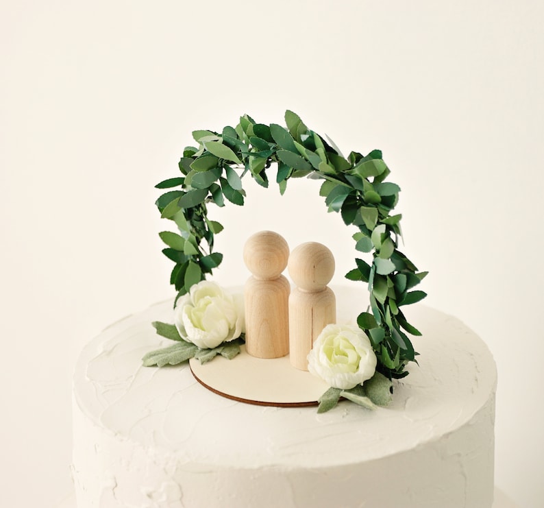 Puede incluir: Un adorno para tarta de boda con dos mu&ntilde;ecos de madera sobre una base de madera, bajo un arco de hojas verdes. Flores blancas y vegetaci&oacute;n se colocan alrededor de la base del arco. La tarta tiene glaseado blanco.