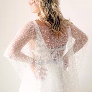 May include: A sheer, white bridal cape with long sleeves, densely covered in sparkling sequins. The cape is worn over a white dress with floral lace detailing. The back of the dress features a bow. The overall look is elegant and ethereal.