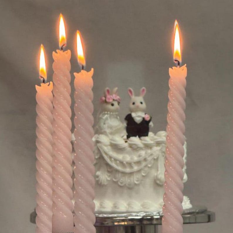 May include: Four tall, light pink spiral taper candles with lit flames stand in front of a white cake topped with two rabbit figurines. One rabbit wears a white dress with a floral crown, and the other wears a black suit with a white bowtie.