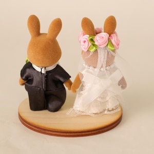 May include: A pair of brown rabbit figurines dressed as a bride and groom. The groom wears a black tuxedo, and the bride has a white dress, veil, and a floral crown. They stand on a round wooden base.