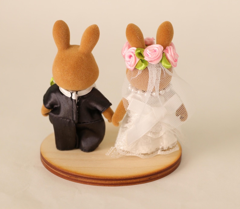 May include: A pair of brown rabbit figurines dressed as a bride and groom. The groom wears a black tuxedo, and the bride has a white dress, veil, and a floral crown. They stand on a round wooden base.