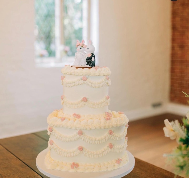 May include: A two-tiered white cake decorated with piped frosting swags and small pink floral accents. The top of the cake features a figurine of two white rabbits, one wearing a white dress and pink bow, the other in a black suit.