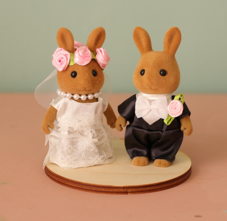 May include: Two brown rabbit figurines dressed as a bride and groom. The bride wears a white dress, veil, and a pink flower crown. The groom is in a black tuxedo with a white bow tie and a pink rose boutonniere. They stand on a wooden base.