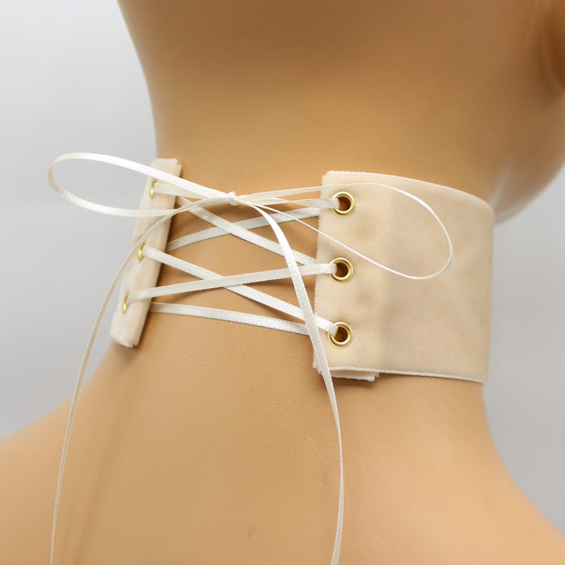 May include: A cream-colored velvet choker with white satin ribbon lacing and gold eyelets. The ribbon is tied in a bow at the back of the neck.