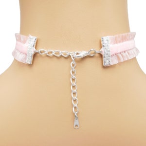 May include: A pink velvet choker with a delicate lace trim and silver chain closure. The choker has two silver rectangular clasps with a textured design.