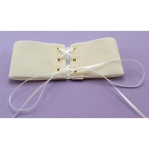 May include: A cream-colored velvet corset with white ribbon lacing and gold eyelets. The corset is tied in the back and the ribbon laces are loose.
