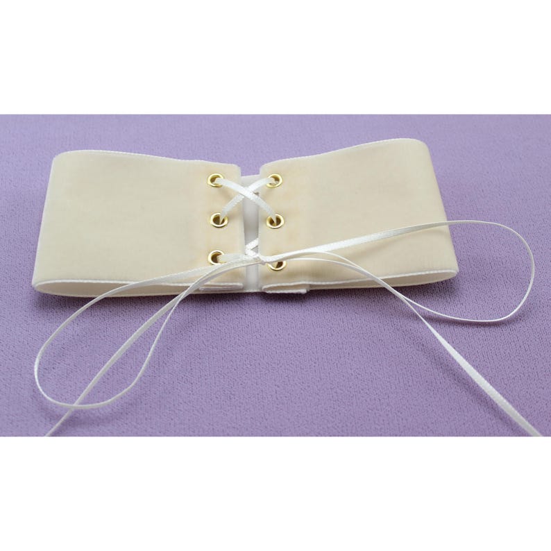 May include: A cream-colored velvet corset with white ribbon lacing and gold eyelets. The corset is tied in the back and the ribbon laces are loose.