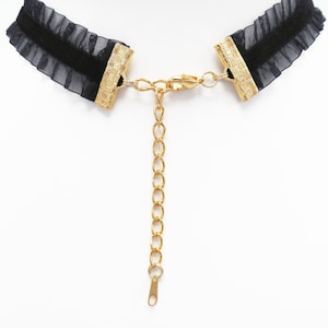 May include: A black choker necklace with a gold chain extender. The choker is made of black mesh fabric with a ruffled edge. The chain extender is made of gold metal and has a lobster clasp.