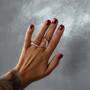May include: A polished silver ring featuring an open, curved design that spans across two fingers. The metal has a smooth, reflective surface with a continuous, fluid shape that creates a wide, arched silhouette across the hand.