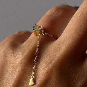 Puede incluir: Un anillo de oro con un pequeño dije en forma de corazón que cuelga de una cadena. El dije en forma de corazón está adornado con pequeñas piedras brillantes. El anillo está en un dedo.