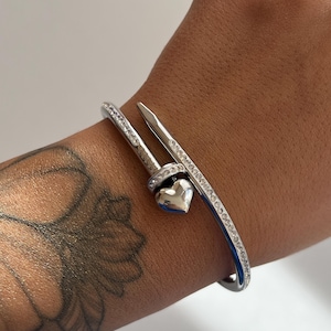 May include: A silver-toned bracelet with a nail-inspired design. The bracelet features a heart-shaped charm and is embellished with small, clear crystals. The bracelet is worn on a wrist with a tattoo.