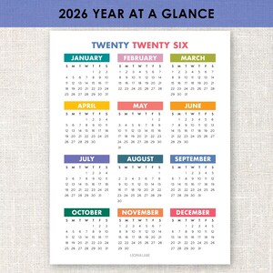 以下が含まれることがあります： 白地の2026年年間カレンダー。各月は異なる色の見出しが付いています。「Twenty Twenty Six」の文字が上部にあります。カレンダーはLion & Lane製です。