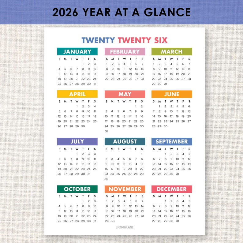 以下が含まれることがあります： 白地の2026年年間カレンダー。各月は異なる色の見出しが付いています。「Twenty Twenty Six」の文字が上部にあります。カレンダーはLion & Lane製です。