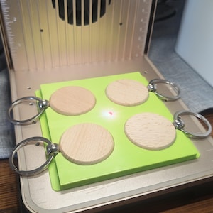 May include: Four round wooden keychains with silver key rings are arranged on a bright green surface. The keychains are inside a gold-colored laser engraving machine. The machine has a fan and a ruler on the side.