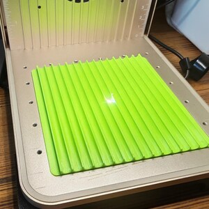 May include: A laser engraving machine with a bright green platform. The machine has a silver metal casing with a fan and a ruler printed on the side. A laser beam is visible on the green platform.