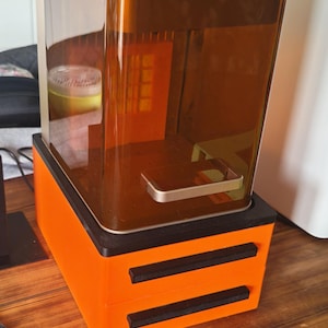 Könnte beinhalten: Ein orangefarbener 3D-Drucker mit einem transparenten, bernsteinfarbenen Harzbehälter. Der Drucker hat eine schwarze Basis und zwei schwarze horizontale Balken auf der Vorderseite. Das Gerät steht auf einer Holzoberfläche.