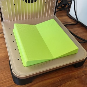 May include: A laser engraving machine with a bright green, folded material inside. The machine has a beige frame with vertical slots and a black base. The material appears to be a foam or rubber. The machine is on a wooden surface.