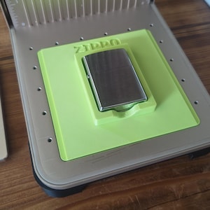 May include: A silver Zippo lighter rests in a lime green holder, set within a gray device. The word "Zippo" is printed on the holder. The device has a ruler-like scale on the side.