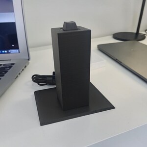 May include: A black charging stand with a rectangular base and a tall, square body. A black cord is attached to the stand. A laptop and tablet are visible in the background.