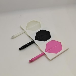 Puede incluir: Un juego de tres plumas para dardos con sus respectivas varillas en blanco, negro y rosa. Cada pluma está unida a una varilla del mismo color, dispuestas en fila sobre una tarjeta rectangular blanca. Los componentes están fabricados en plástico.